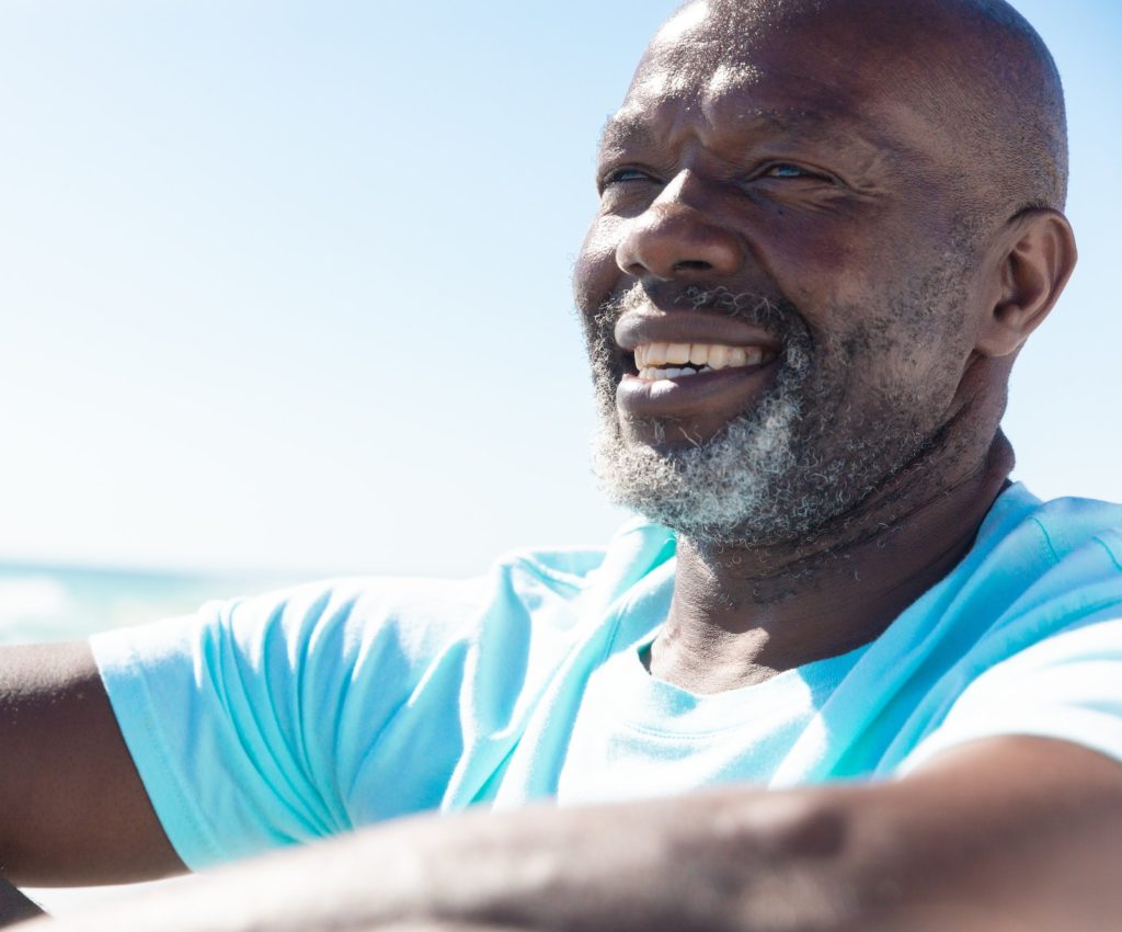 Smiling african american retired senior man with white beard sitting at beach on sunny day