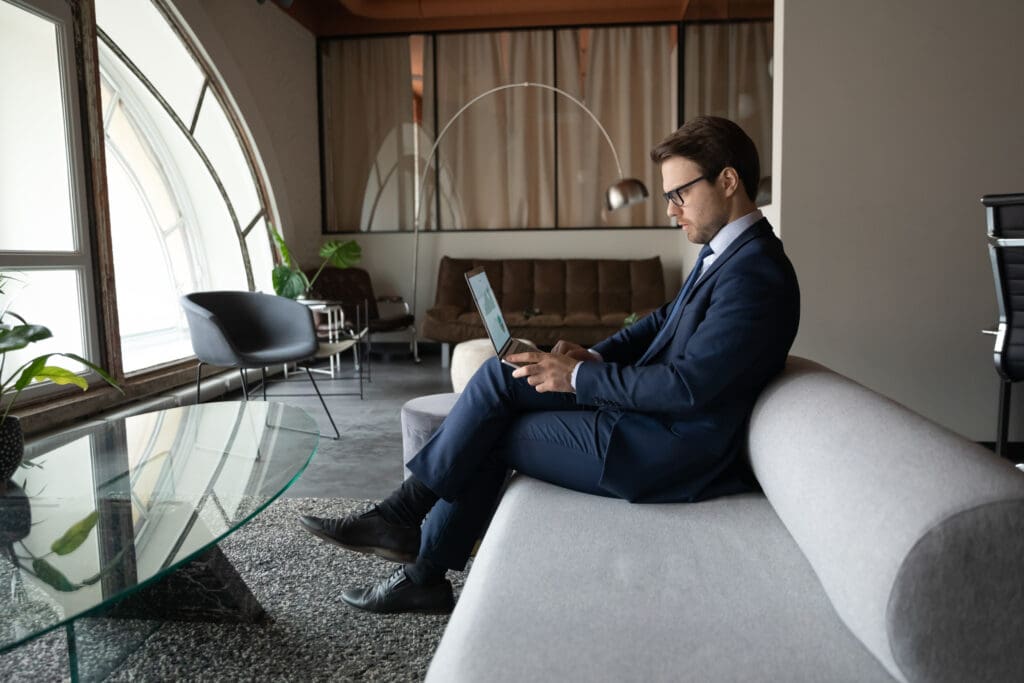 A bespectacled businessman in a dark blue suit sits on a light grey sofa in a modern, upscale lounge with an arched window, working on a laptop that displays financial charts and graphs.