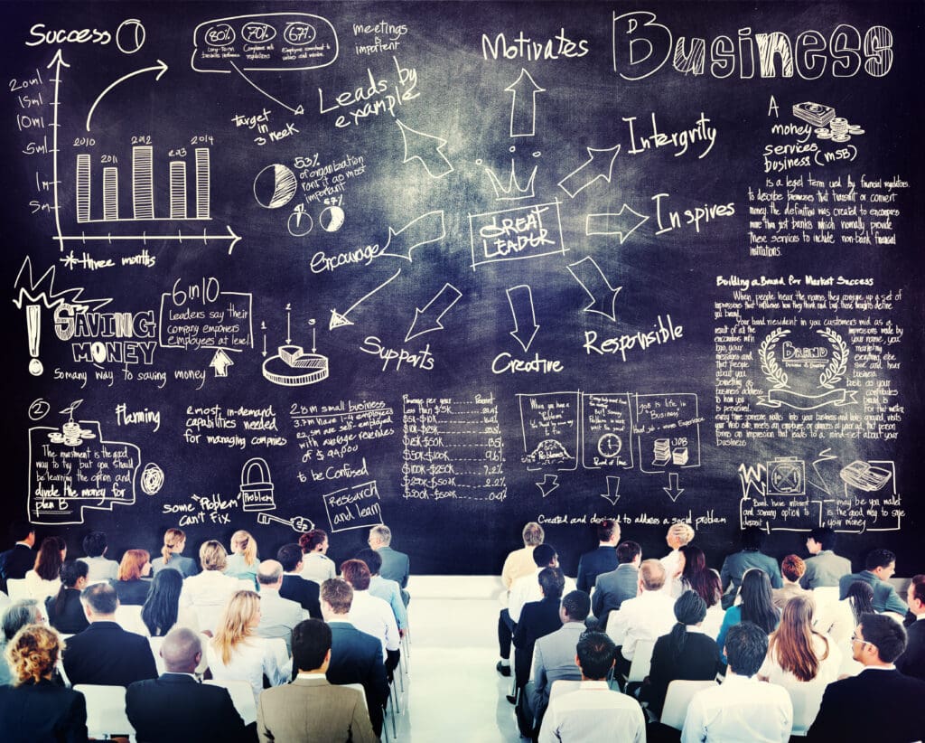 Rows of business professionals sit with their backs to the camera, facing a dark chalkboard densely filled with white hand-drawn business sketches, including bar graphs, pie charts, arrows, and terms such as 'Success,' 'Great Leader,' 'Saving Money,' and 'Brand.'