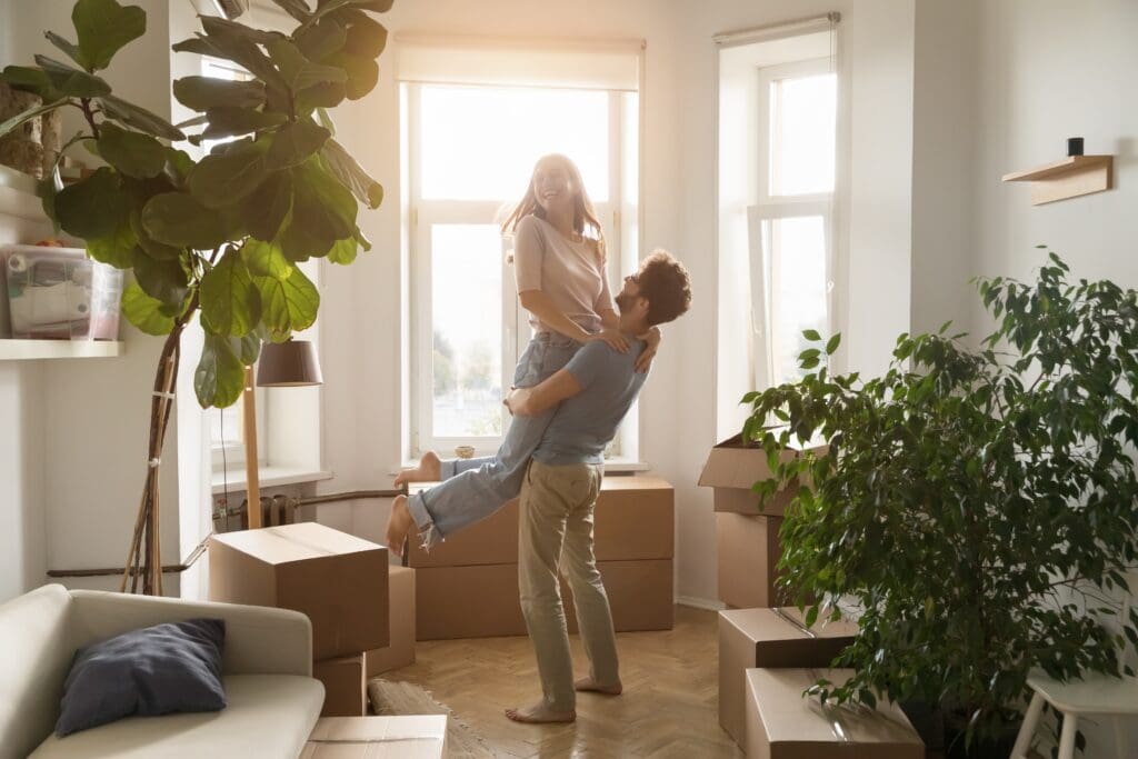 A young couple celebrates moving into their new home, with the man lifting the smiling woman in a sunlit living room surrounded by cardboard moving boxes and houseplants.