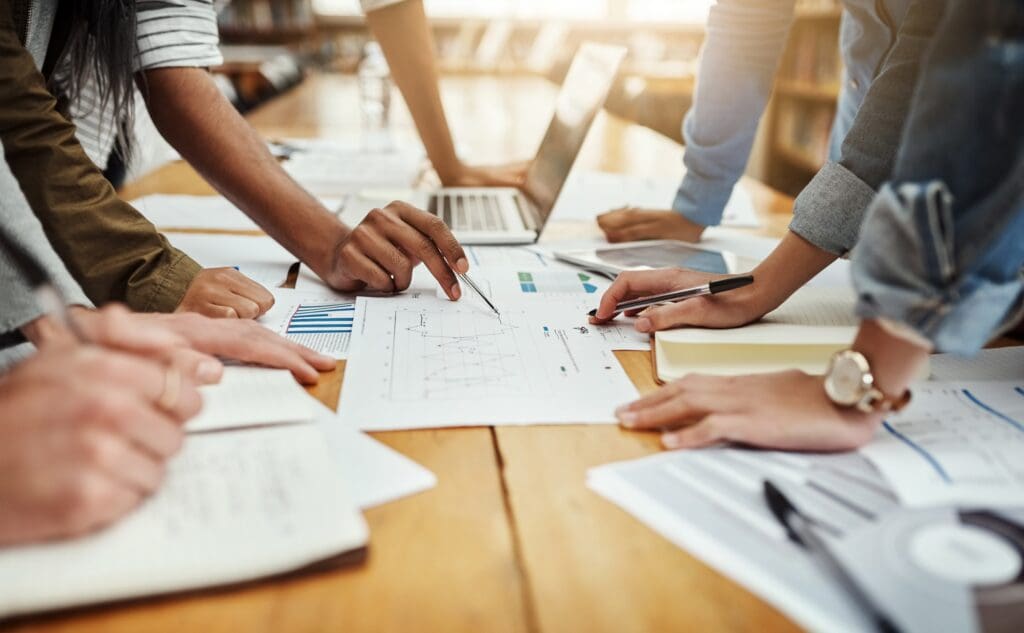 Close-up of multiple hands gathered around a wooden table strewn with financial reports, bar charts, and line graphs, as team members point at data with pens alongside a laptop, tablet, and notebooks in a sunlit room.