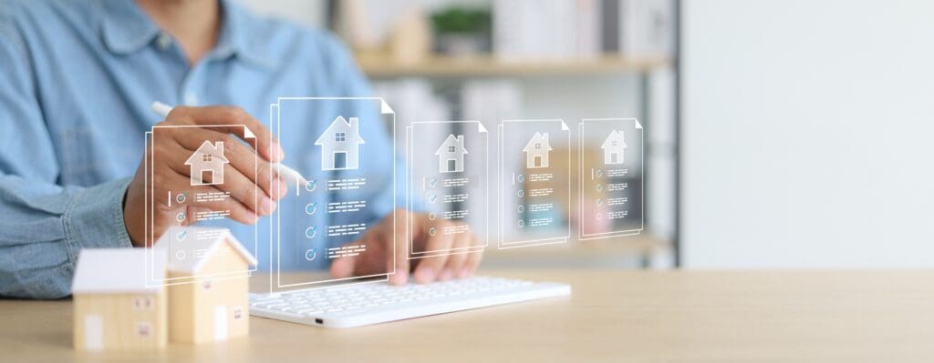 A man in a blue shirt holds a stylus over a white keyboard, with wooden toy house models on the desk and a futuristic overlay of five translucent document cards, each showing a house icon and a checklist of items to compare.