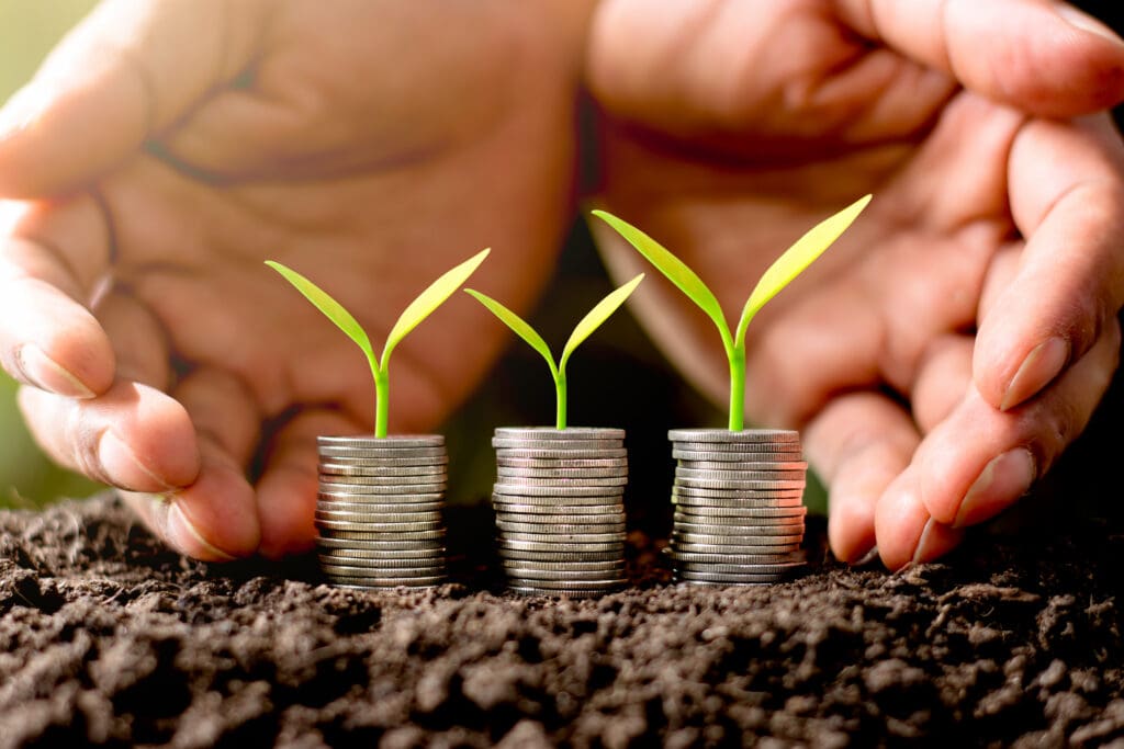 Hands cradling three rising stacks of coins topped with green seedlings, growing in soil