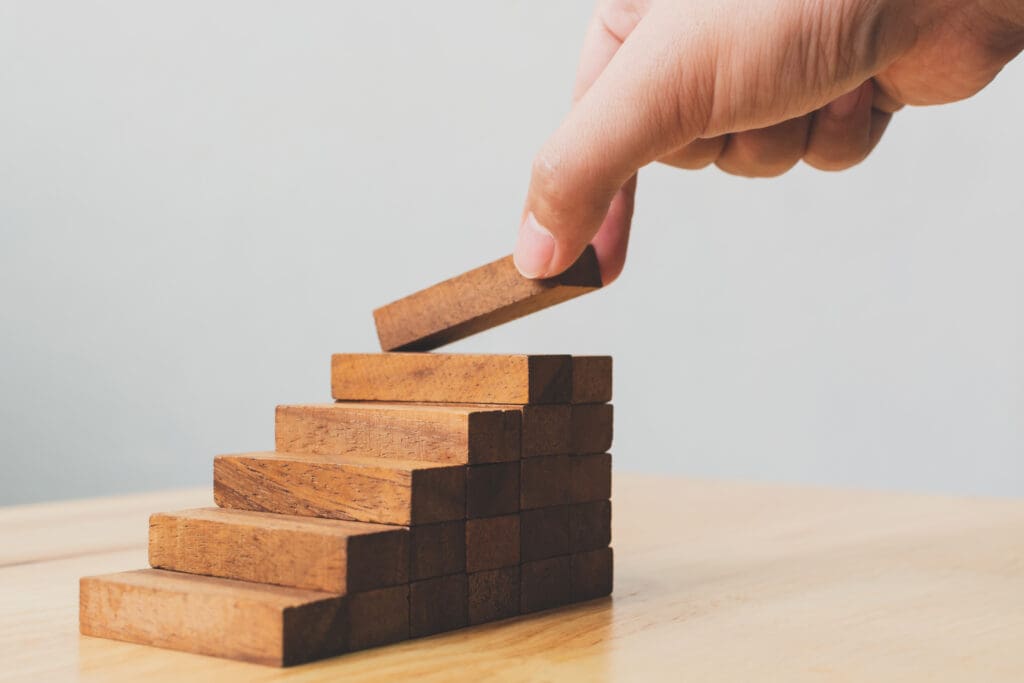 A hand placing a wooden block on top of a rising staircase built from stacked wooden blocks, set against a light gray background